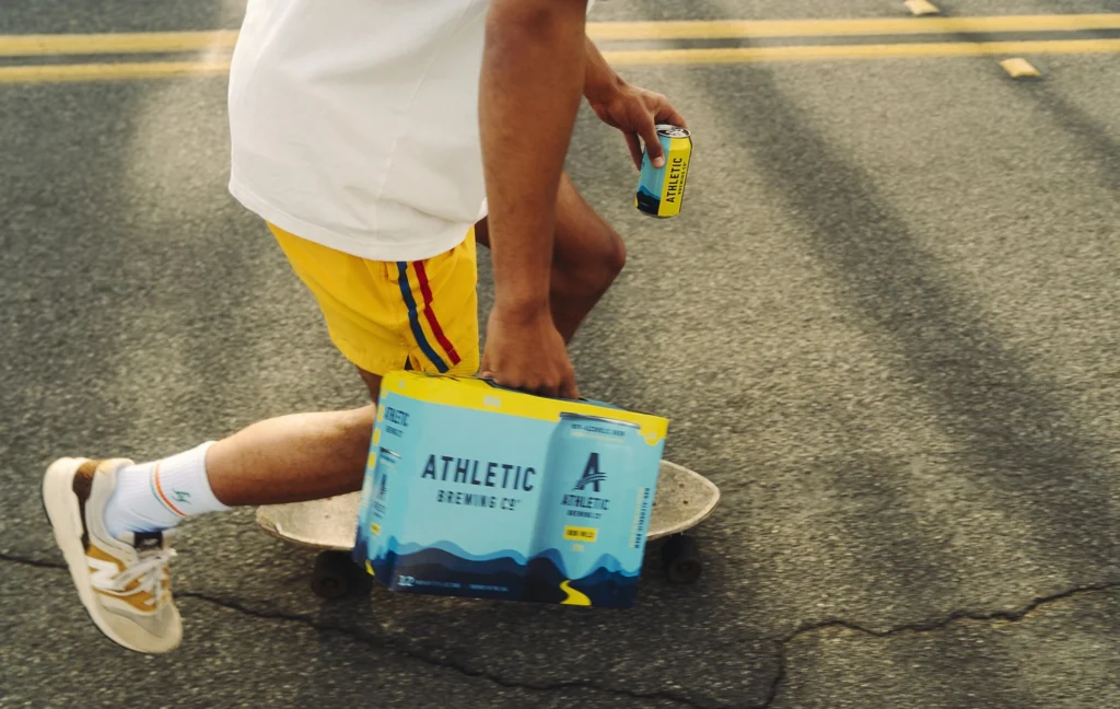 a man on a skateboard carrying a case of Athletic Brewing beer.