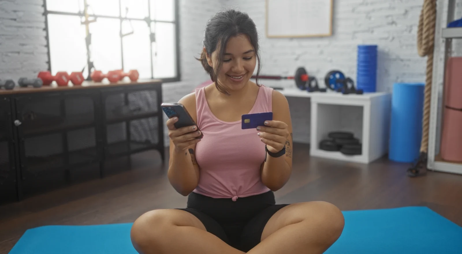 A young hispanic woman in a gym uses her smartphone and credit card while sitting on a blue mat