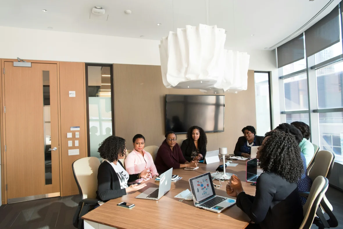 A group of people, including several women, sitting around a conference table with laptops, having a meeting in a modern office with large windows and a TV on the wall.