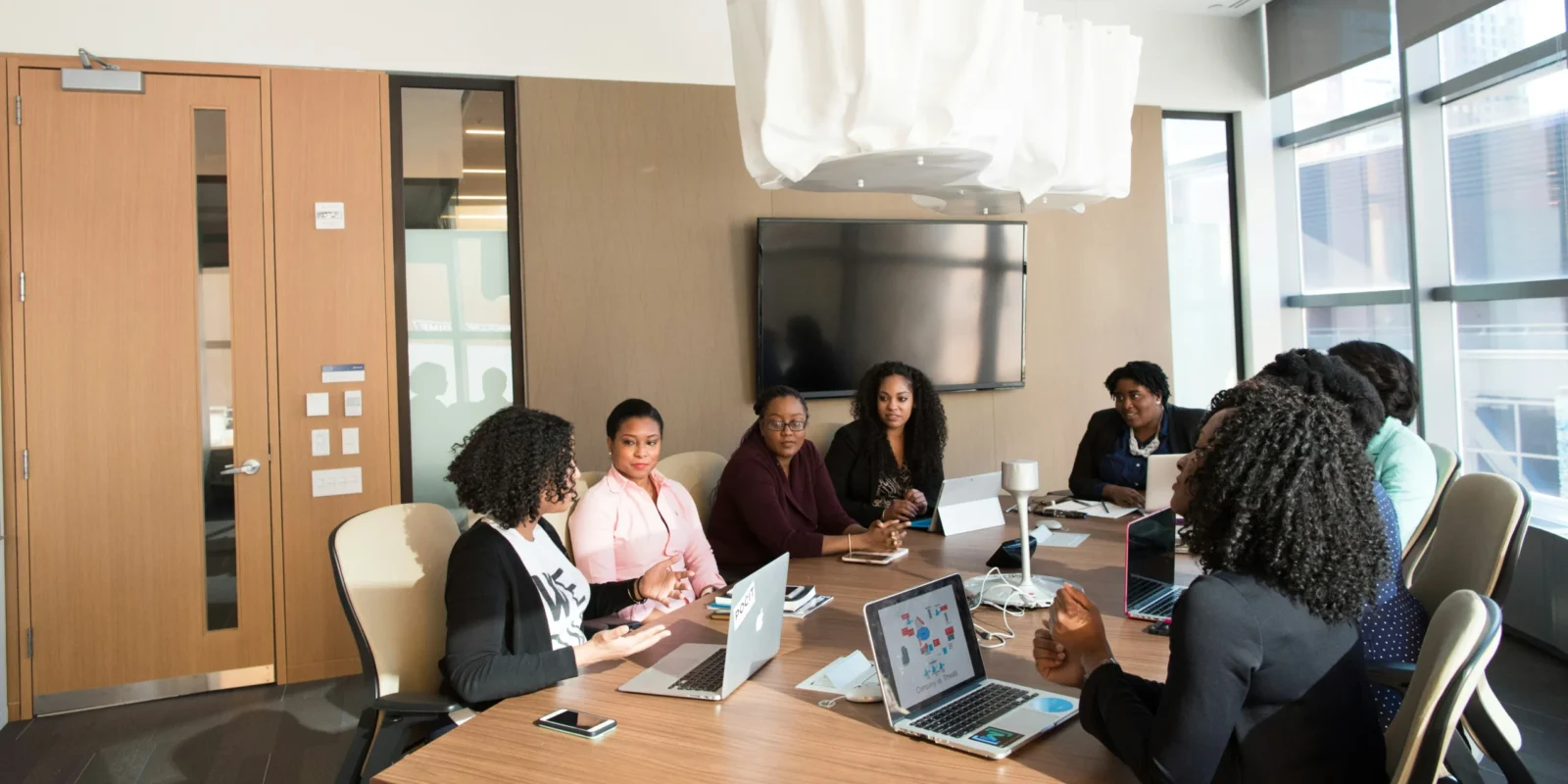 A group of people, including several women, sitting around a conference table with laptops, having a meeting in a modern office with large windows and a TV on the wall.