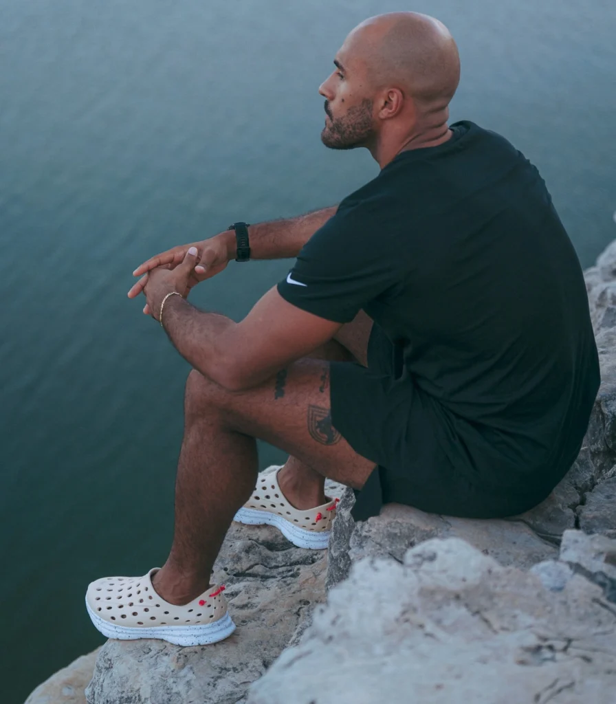 man wears Kane shoes while sitting on a rocky ledge next to a body of water