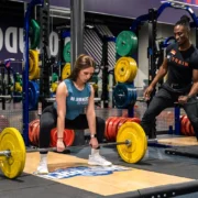 woman picks a barbell up off the ground next to her personal trainer