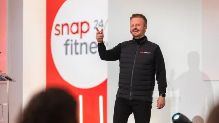 a man on stage wearing a Snap Fitness vest in front of a Snap Fitness sign.