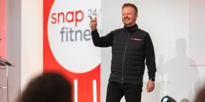 a man on stage wearing a Snap Fitness vest in front of a Snap Fitness sign.