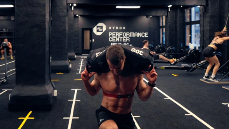 man carries a sandbag inside a Hyrox training club