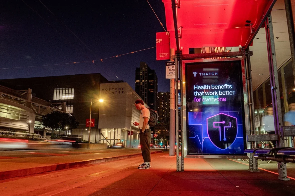 A man standing outside at night near a Thatch billboard.
