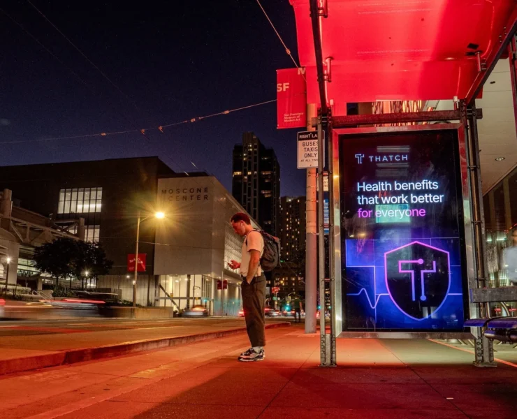 A man standing outside at night near a Thatch billboard.