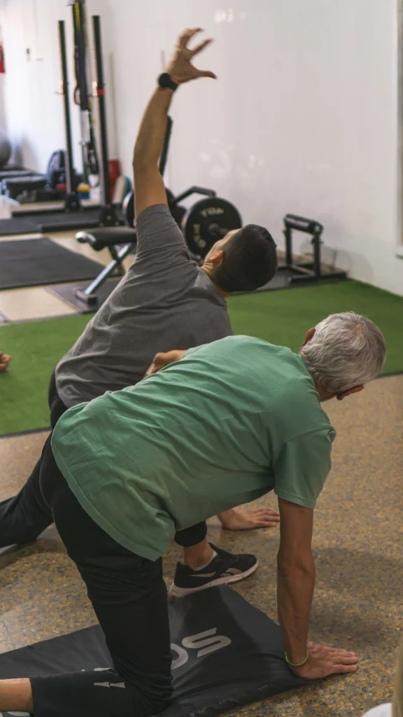 An older gentleman working out with a trainer during yoga on a mat.