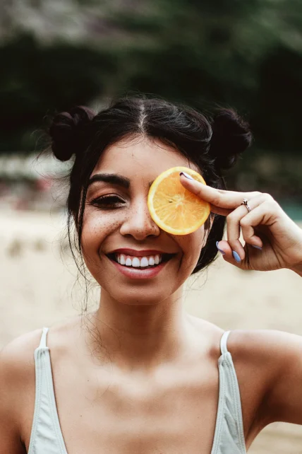 A young woman smiling and holding an orange slice over her left eye.