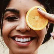 A young woman smiling and holding an orange slice over her left eye.