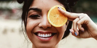 A young woman smiling and holding an orange slice over her left eye.