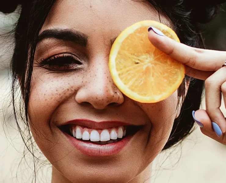 A young woman smiling and holding an orange slice over her left eye.