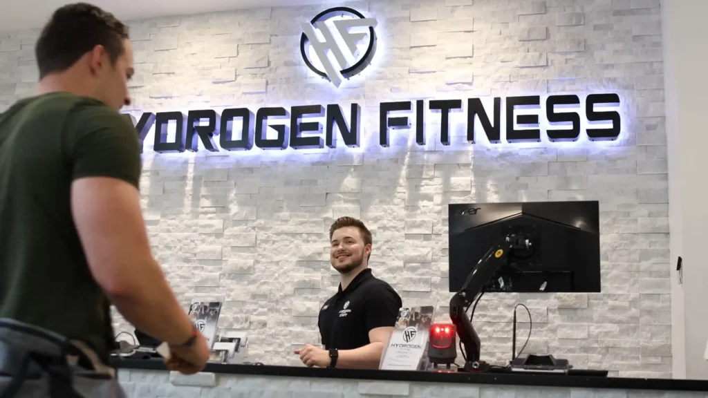 The front desk at Hydrogen Fitness with a smiling man behind it greeting another man.