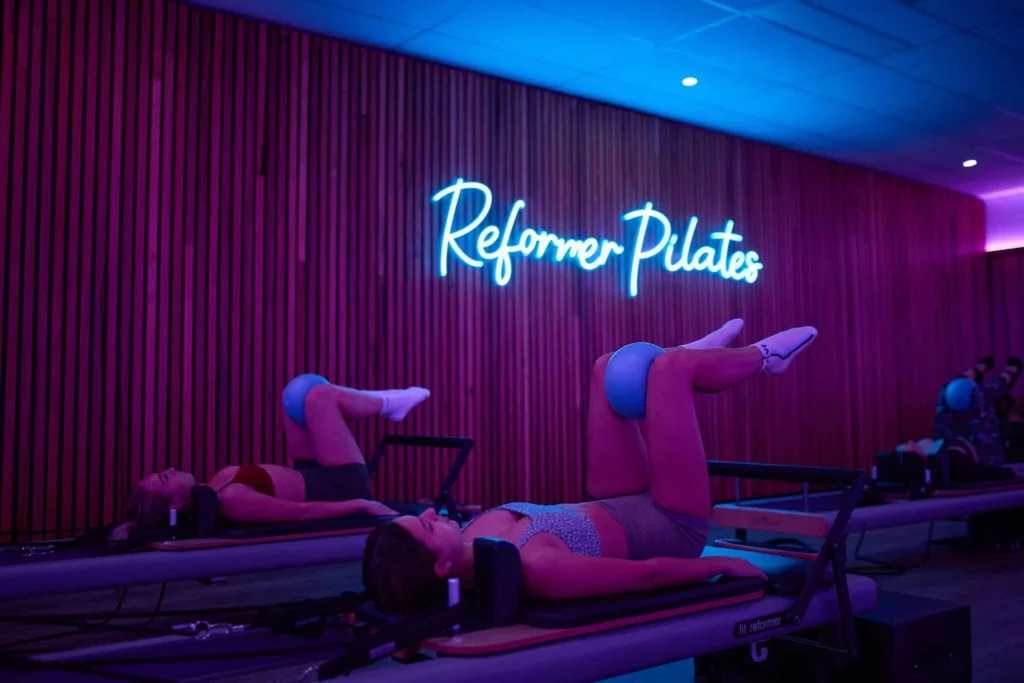 Women working out at Genesis Health + Fitness under a 'Reformer Pilates' LED sign.