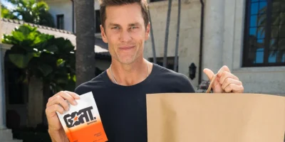 Tom Brady outdoors holding a pack of GOAT Gummies and a GOPUFF bag.