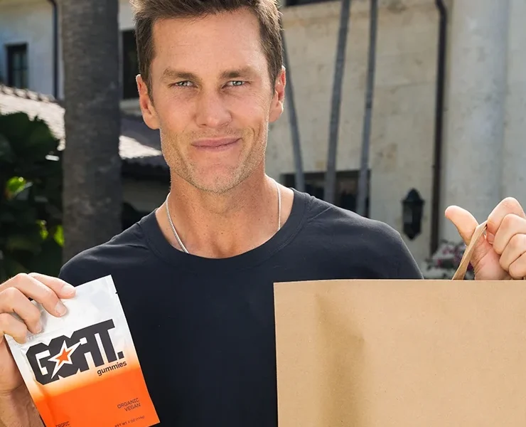 Tom Brady outdoors holding a pack of GOAT Gummies and a GOPUFF bag.