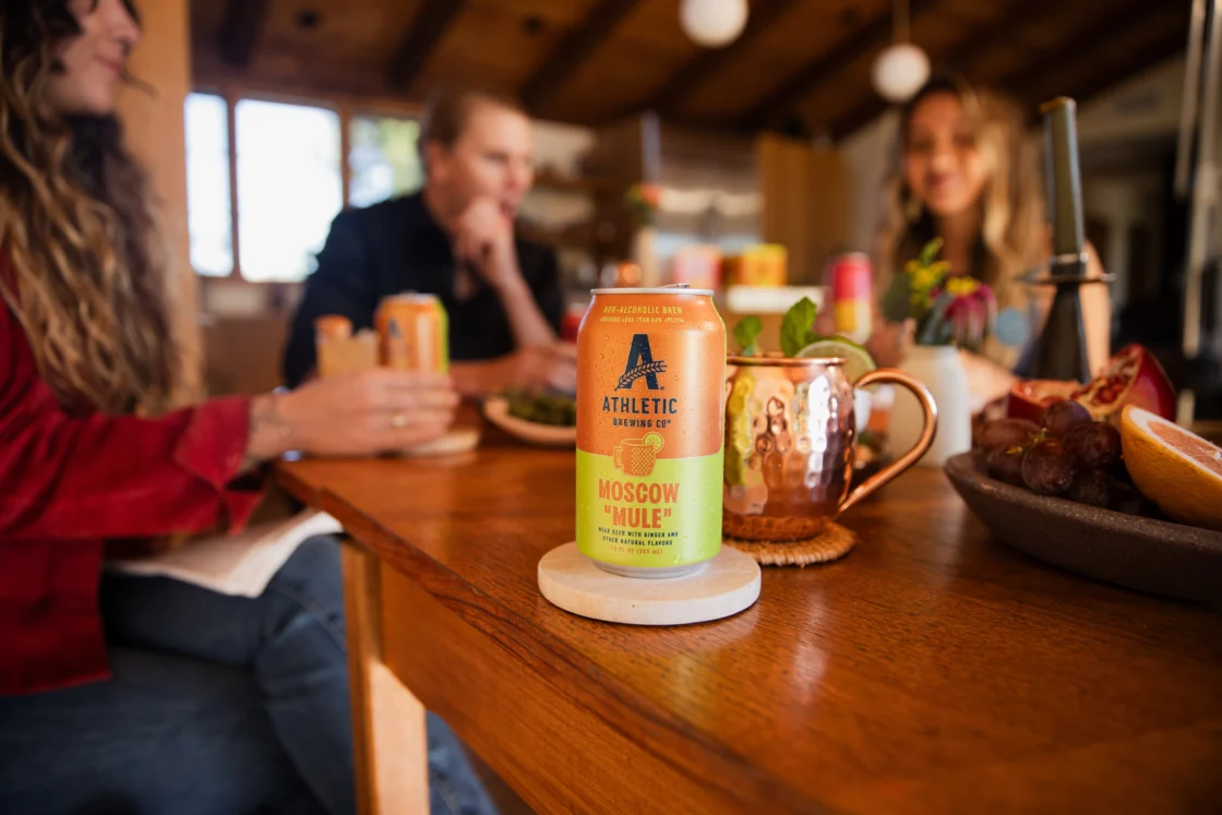 Athletic Brewing Moscow Mule non-alcoholic beer on a dining table with friends gathered, highlighting alcohol-free cocktail-inspired brews