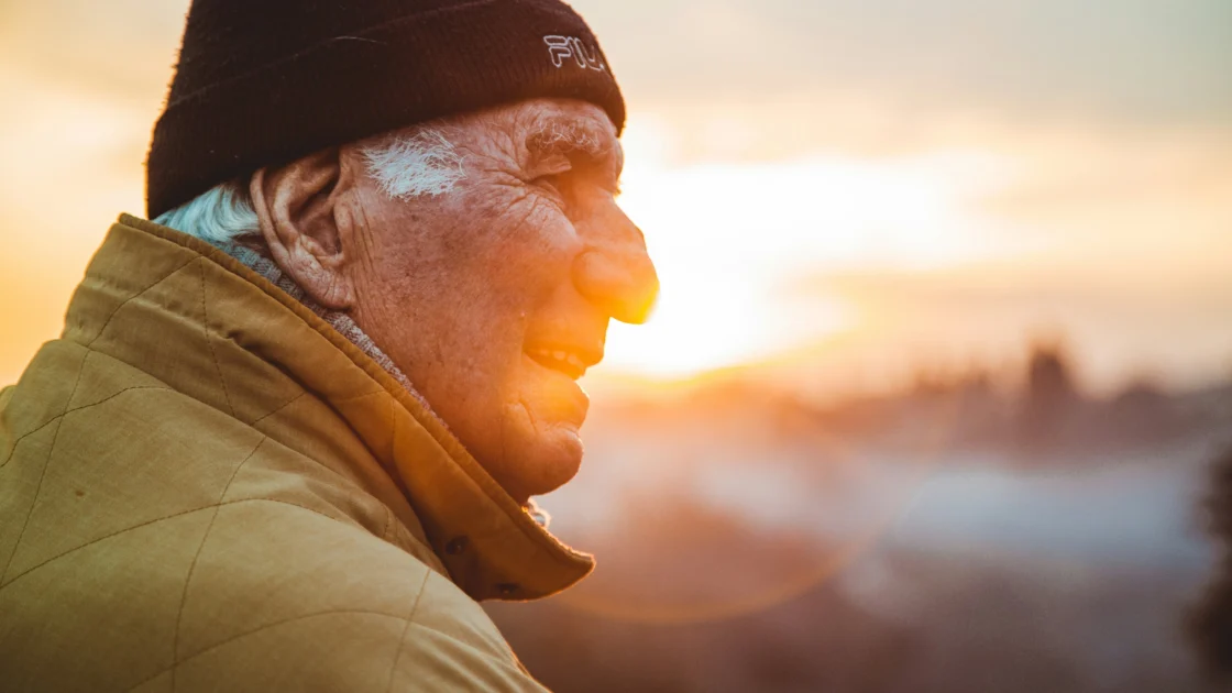 Elderly man in a beanie and jacket gazes toward the sunrise, his face lit by warm morning light against a softly blurred landscape.