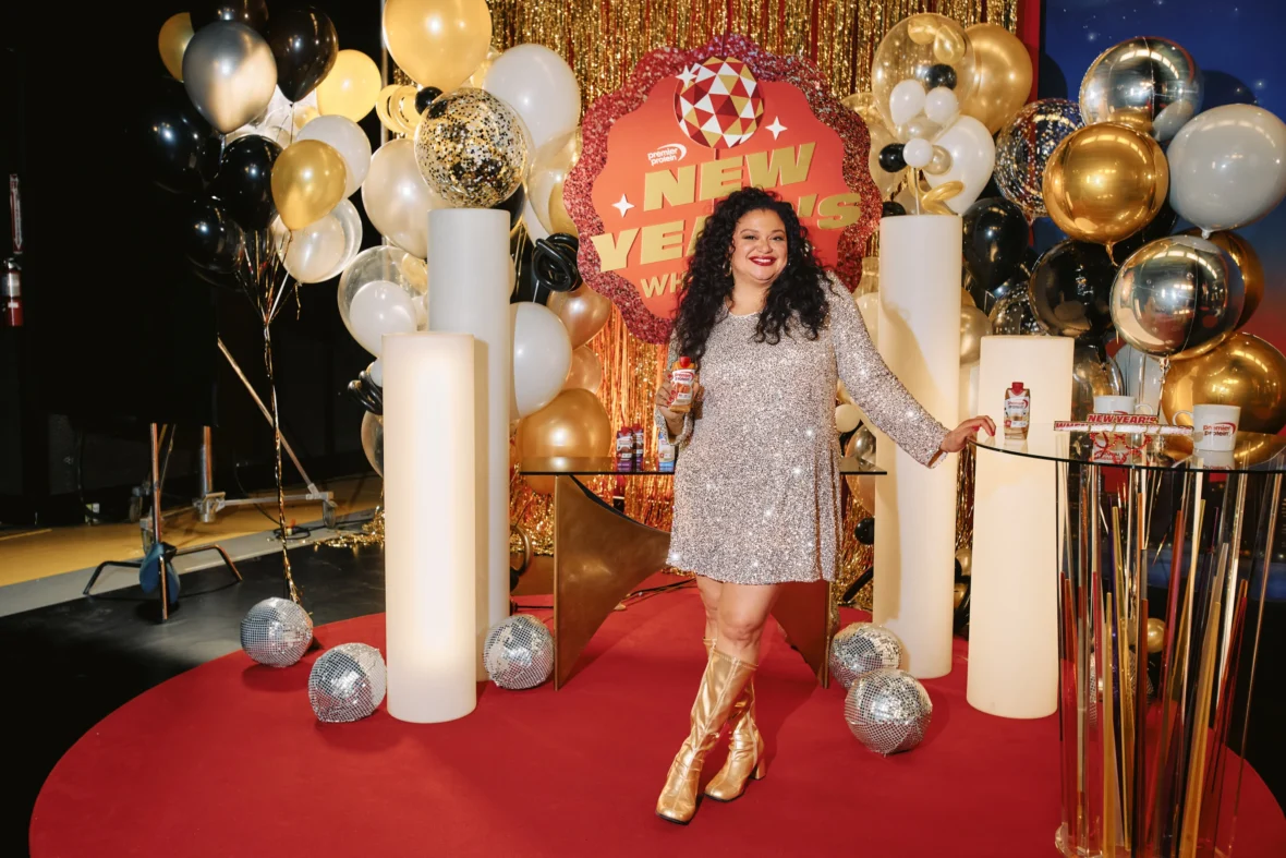 Michelle Buteau is seen in a silver sequined dress and gold boots holds a Premier Protein shake while posing on a festive New Year–themed set with balloons and branded signage.