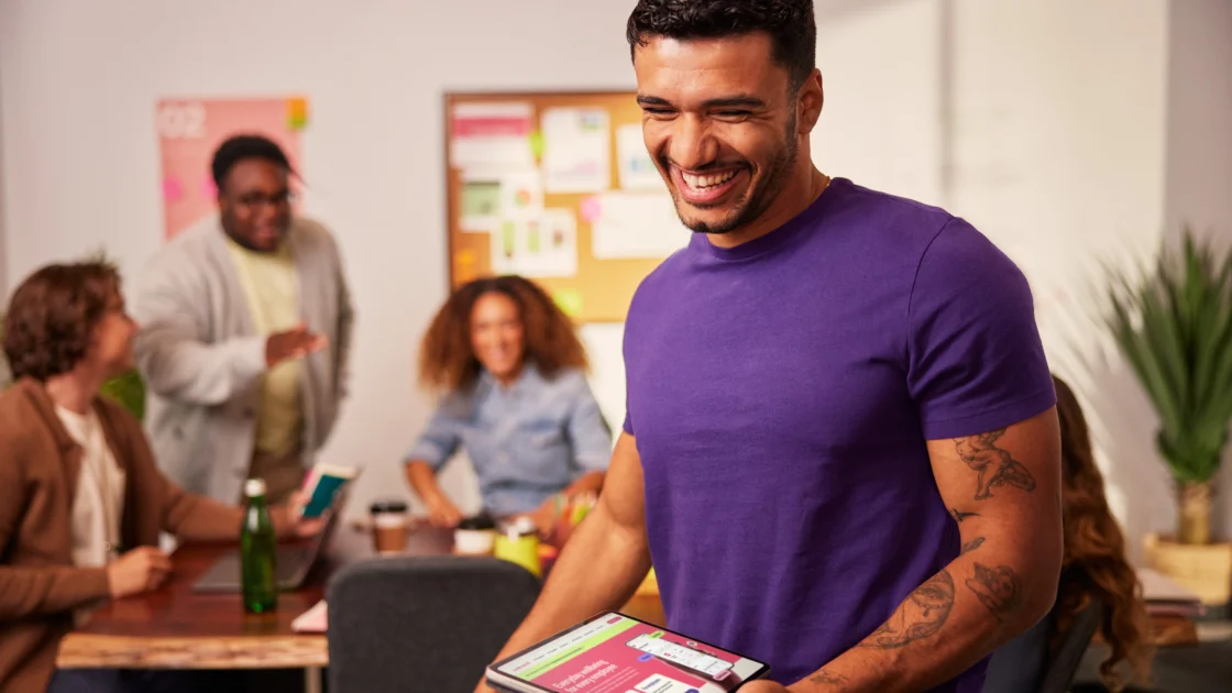 Smiling employee holding a tablet with a wellness app open while colleagues chat in a modern office setting.