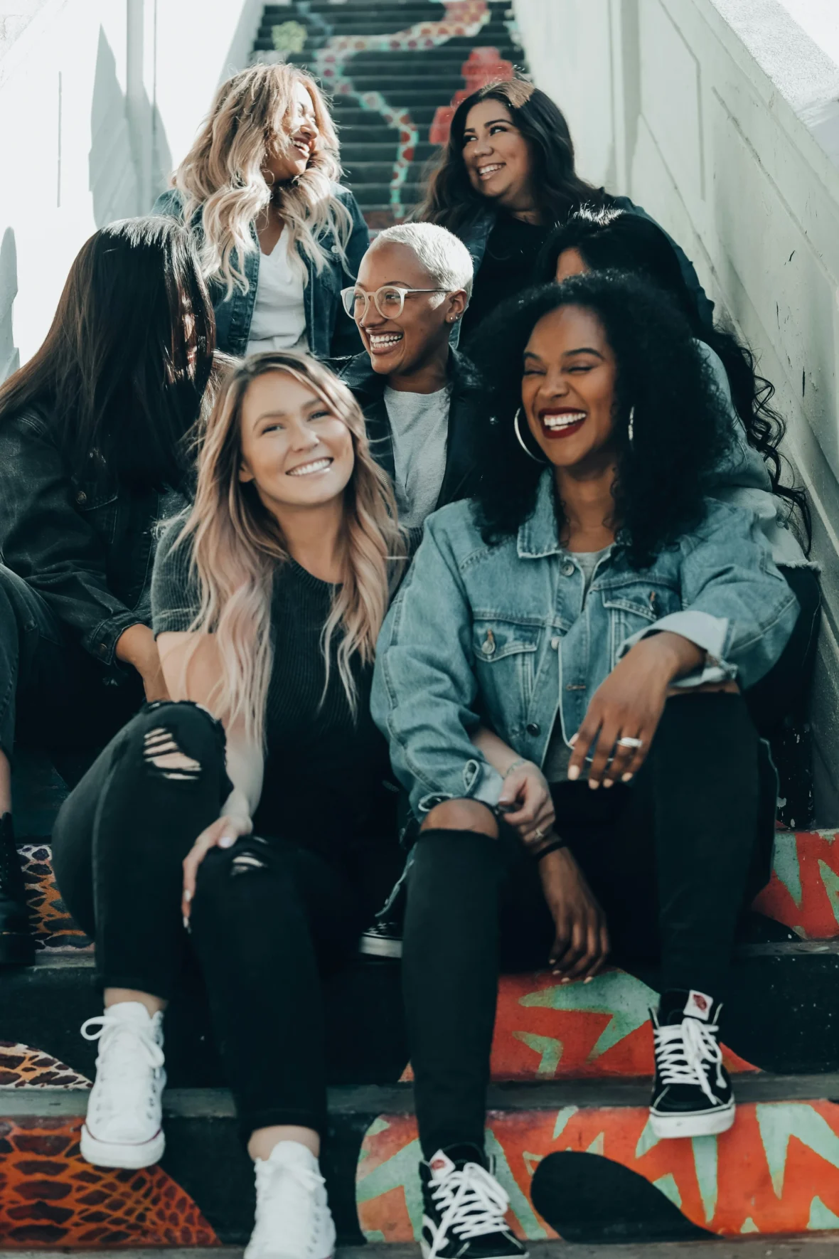A diverse group of seven women sit together on colorful painted stairs, laughing and smiling, dressed casually in denim jackets, T-shirts and sneakers.