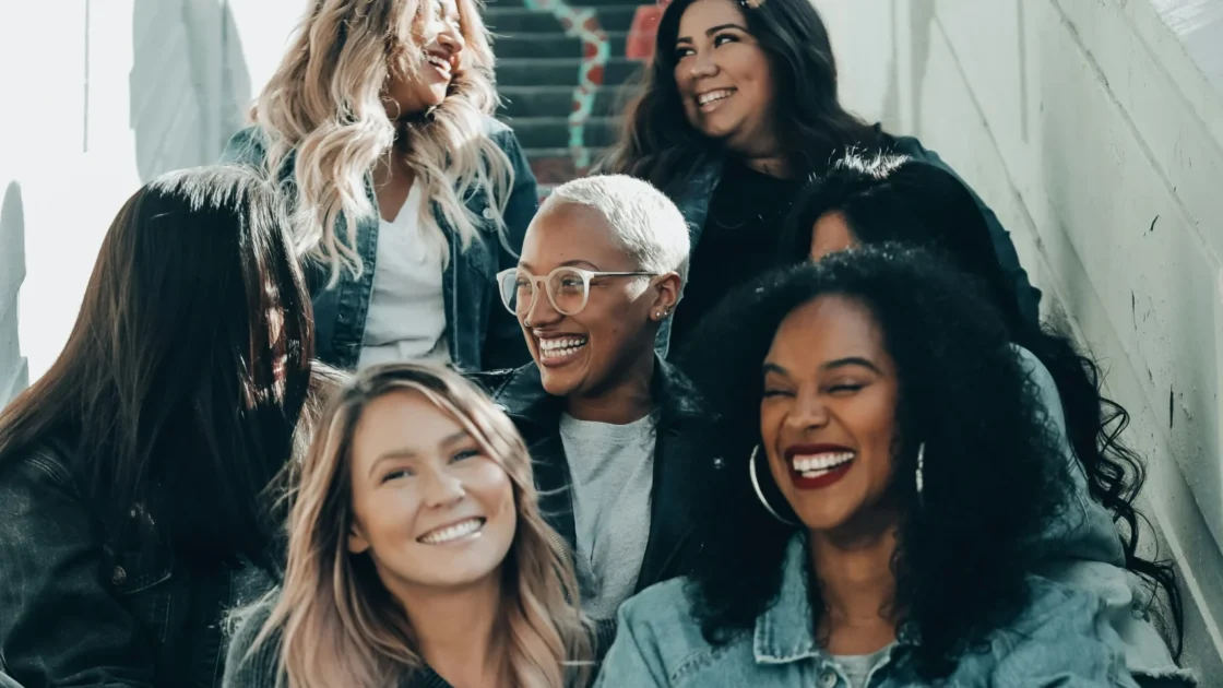 A diverse group of seven women sit together on colorful painted stairs, laughing and smiling, dressed casually in denim jackets, T-shirts and sneakers.