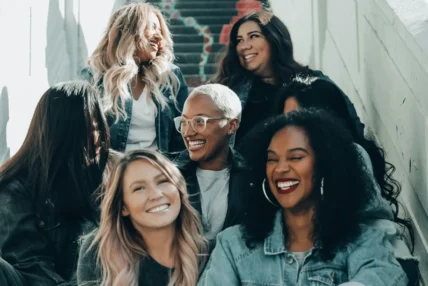 A diverse group of seven women sit together on colorful painted stairs, laughing and smiling, dressed casually in denim jackets, T-shirts and sneakers.