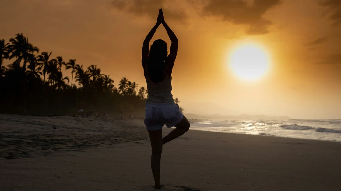 A silhouetted person balances in tree pose on a beach at sunset, with palms in prayer overhead as the sun sets over the ocean.
