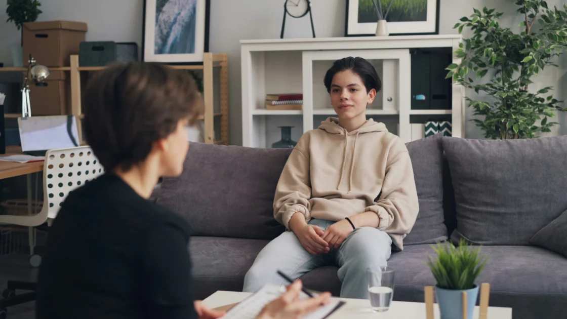 A young person sits on a couch speaking with a therapist during a counseling session in a home-like office setting.