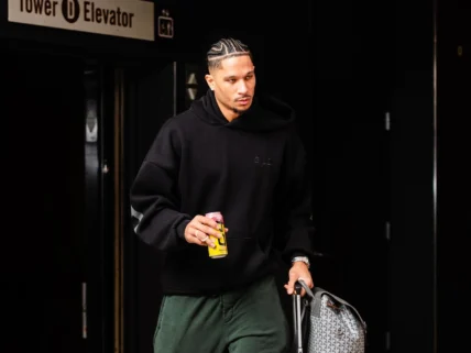 Knicks guard Josh Hart walking with rolling suitcase and backpack while holding an energy drink, dressed in black hoodie and green pants in a modern indoor setting