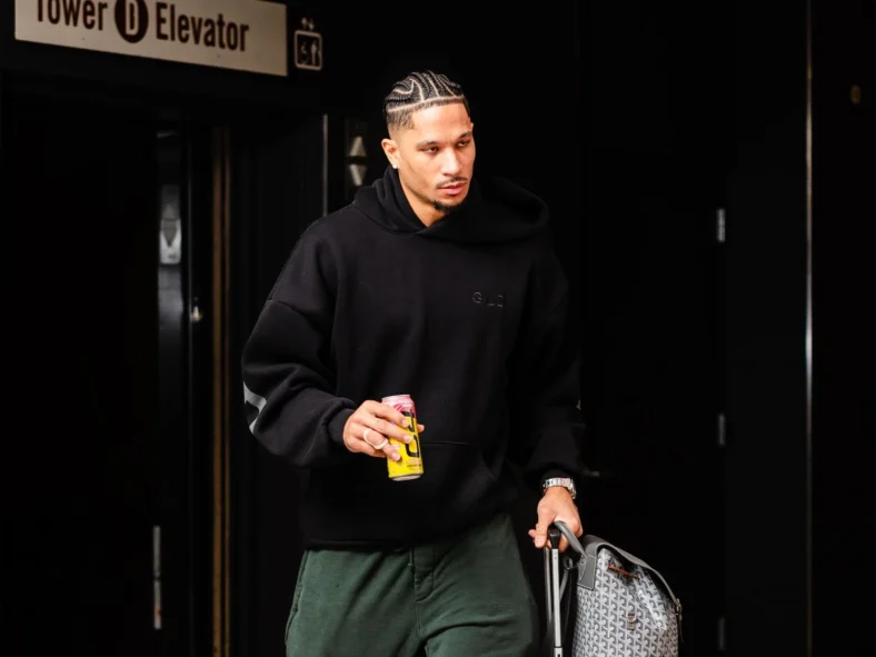 Knicks guard Josh Hart walking with rolling suitcase and backpack while holding an energy drink, dressed in black hoodie and green pants in a modern indoor setting