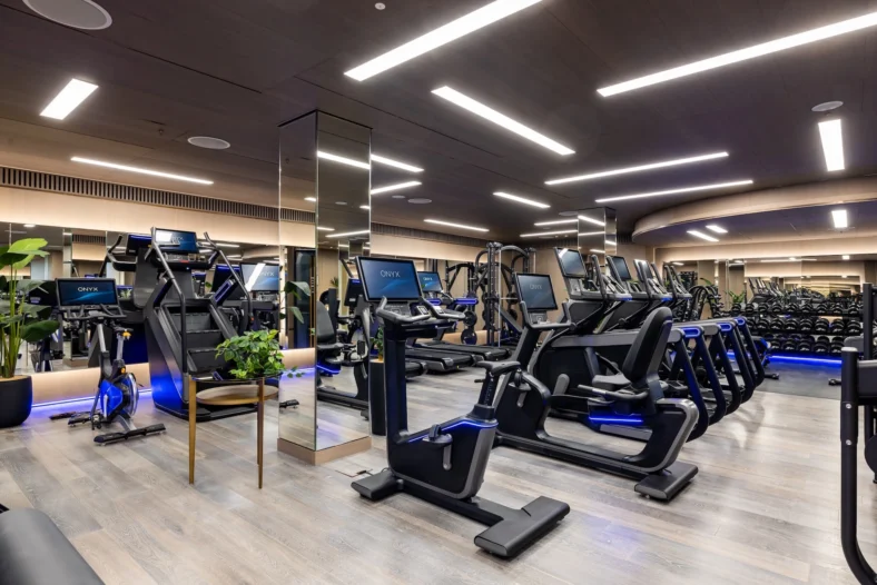 Modern luxury hotel gym with ONYX cardio machines, treadmills, bikes and dumbbell rack, mirrored walls, wood flooring and LED lighting.